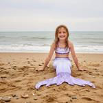 Load image into Gallery viewer, Young girl in a mermaid tail sitting on a beach with ocean in the background