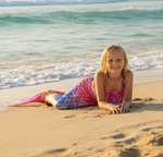 Load image into Gallery viewer, Young girl in a mermaid tail lying on the beach with ocean waves in the background