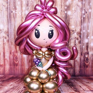 Balloon sculpture of a mermaid with pink hair and gold body against a wooden background