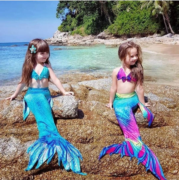 Two children in mermaid tails on a rocky beach with ocean and trees in the background.