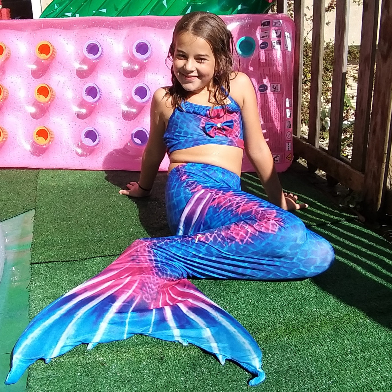 Smiling girl on pool deck wearing the striking Rich Blue & Dark Pink Mermaid Tail with added dorsal fins. Complete with matching material halterneck bikini with cute embellished bow. Part of our luxury range, this tail has a high quality side zip. Mini Mermaid Tails