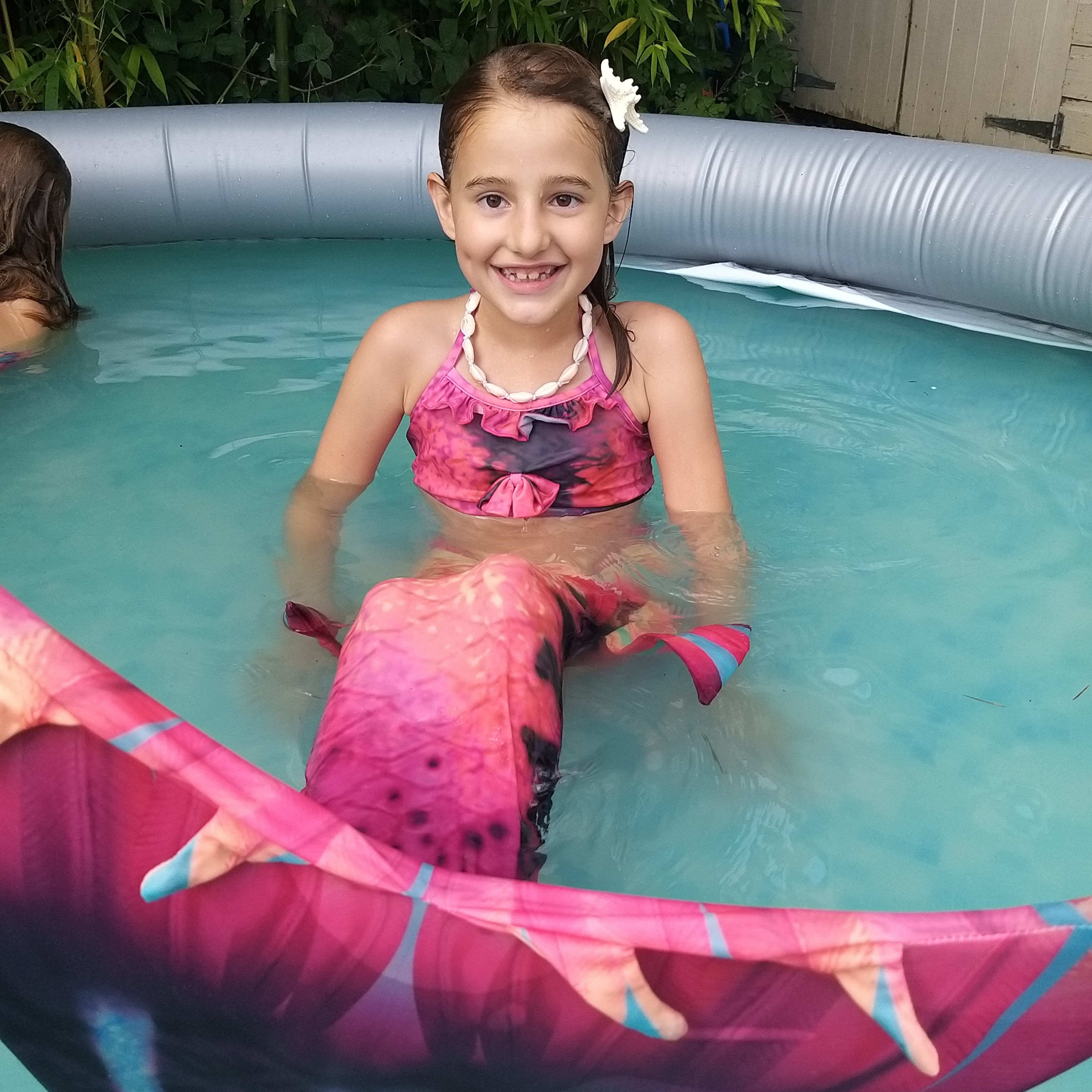 Smiling girl in pool wearing the unusual Pink & Black Mermaid Tail with added dorsal fins. Complete with matching material halter-neck bikini with cute embellished bow.  Part of our luxury range, this tail has a high quality side zip. Mini Mermaid Tails