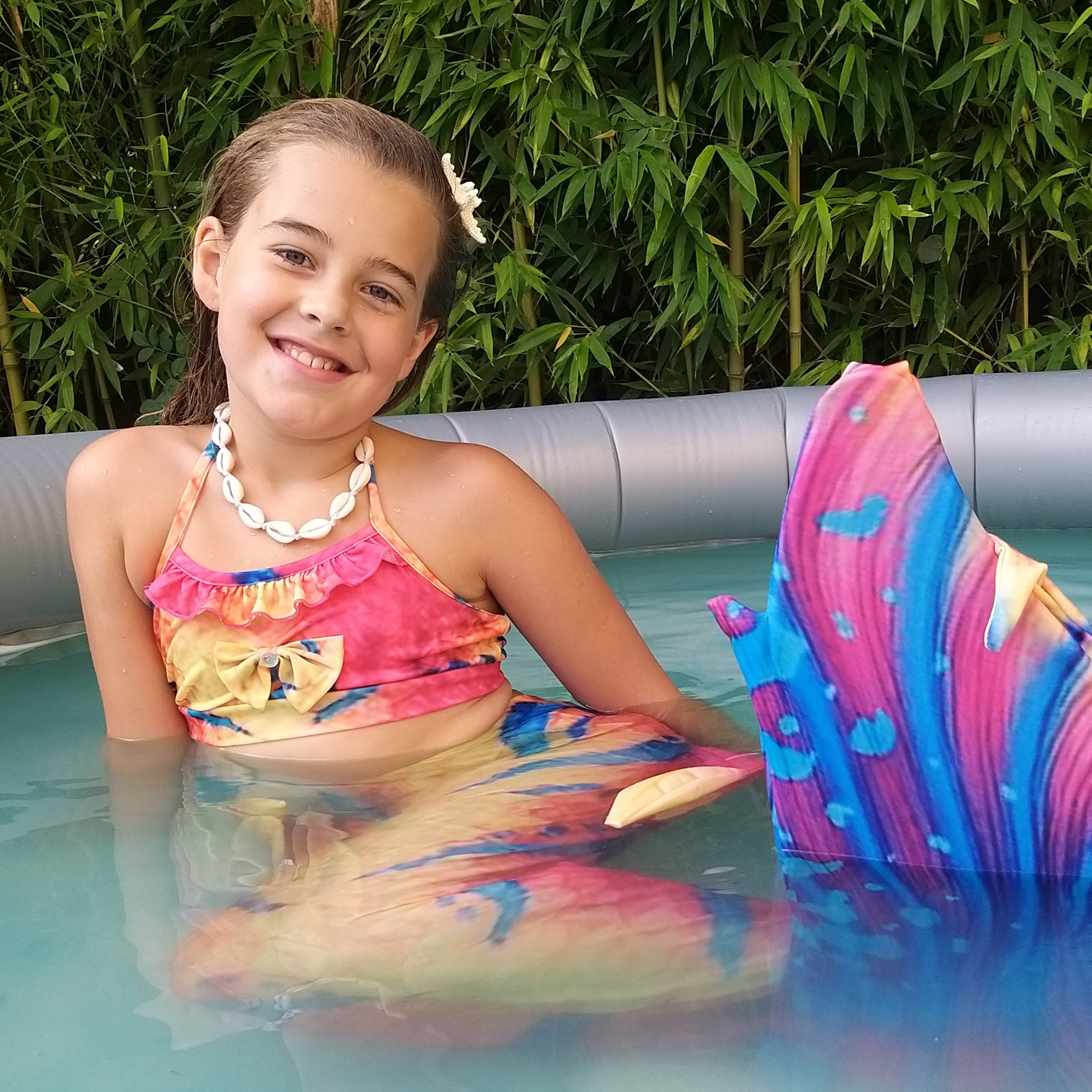Girl in pool wearing the adorable Orange, Yellow & Blue Mermaid Tail with added dorsal fins with matching material halter-neck bikini with cute embellished bow. Part of our luxury range, this tail has a high quality side zip. Mini Mermaid Tails