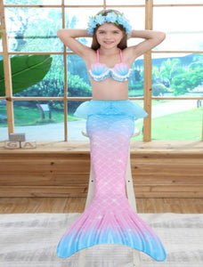 Smiling girl wearing the intricate, slightly padded shell like bikini top with a printed pearl mermaid tail with a netted peplum.  This tail has no fastenings at the bottom so the monofin can be taken on and off easily without stepping out of the whole tail. Mini Mermaid Tails