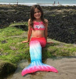 Load image into Gallery viewer, Girl sitting on the beach with her Pink &amp; Blue Mermaid Tail with matching material embellished bow halterneck bikini. Part of our more affordable range, this tail has poppers at the bottom. The monofin can be taken on and off easily without stepping out of the whole tail. Mini Mermaid Tails
