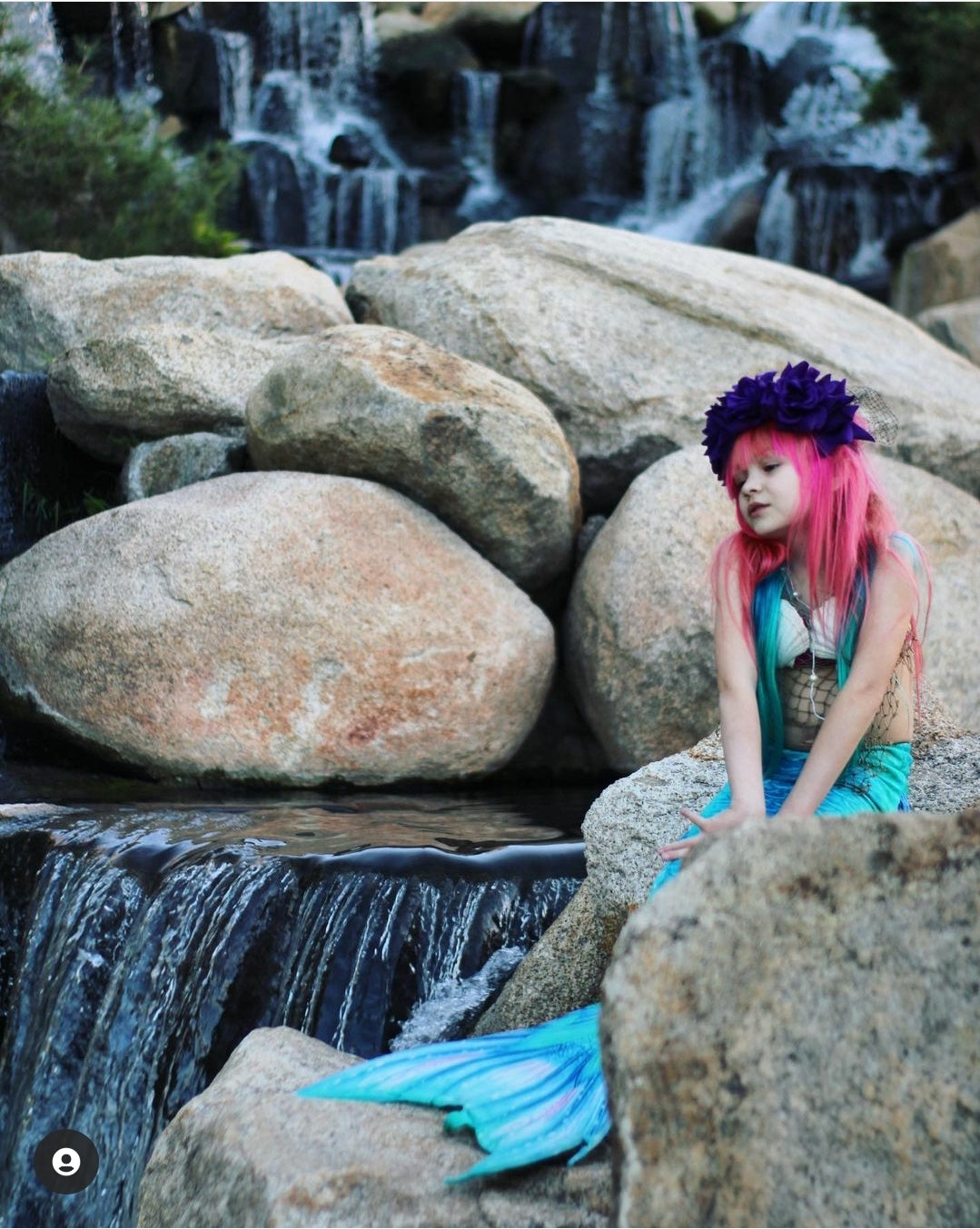 Sweet little girl mermaid with red hair and purple flowers on her head sitting on rocks by a waterfall and wearing the Ocean Blue double frill bikini with matching vibrant mermaid tail. Part of our luxury range, this tail has a high quality side zip. Mini Mermaid Tails