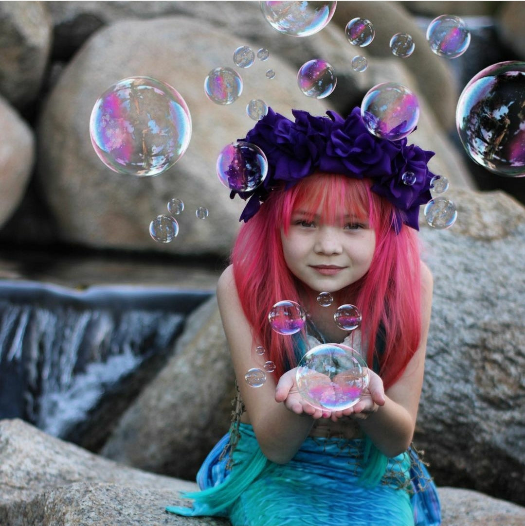 Sweet little girl mermaid with red hair and purple flowers on her head, holding bubbles and wearing the Ocean Blue double frill bikini with matching vibrant mermaid tail. Part of our luxury range, this tail has a high quality side zip. Mini Mermaid Tails